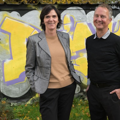 Sigrid Schule und André Eigenbrod, die neuen Leiter der Kreaschule in Bergisch Gladbach, stehen vor einer mit gelb-lilanen Graffitis verzierten Wand.