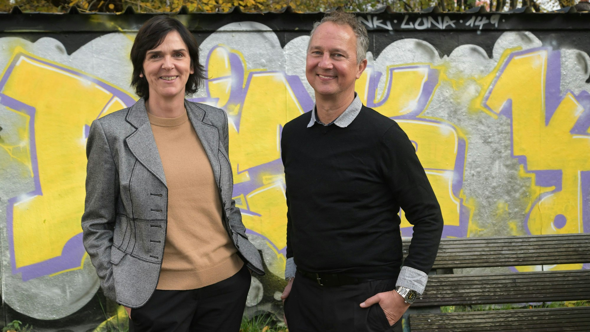Sigrid Schule und André Eigenbrod, die neuen Leiter der Kreaschule in Bergisch Gladbach, stehen vor einer mit gelb-lilanen Graffitis verzierten Wand.