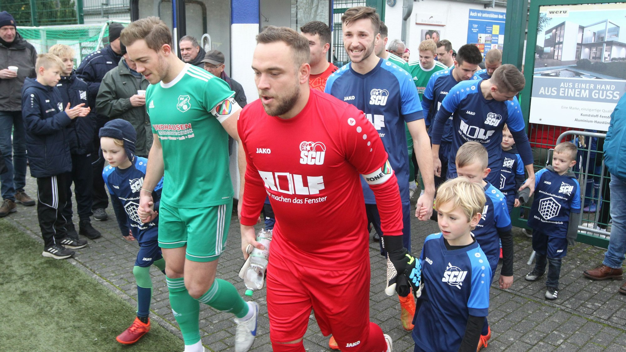 Keeper Fabio Milicki (vorne) vom SC Uckerath und Fabian Ehrenstein (links) vom SV Leuscheid betreten den Kunstrasenplatz in Uckerath mit Kindern an der Hand. Sie tragen die Kapitänsbinde am linken Oberarm mit einem regenbogenfarbigen Herz und der Aufschrift „One Love“.