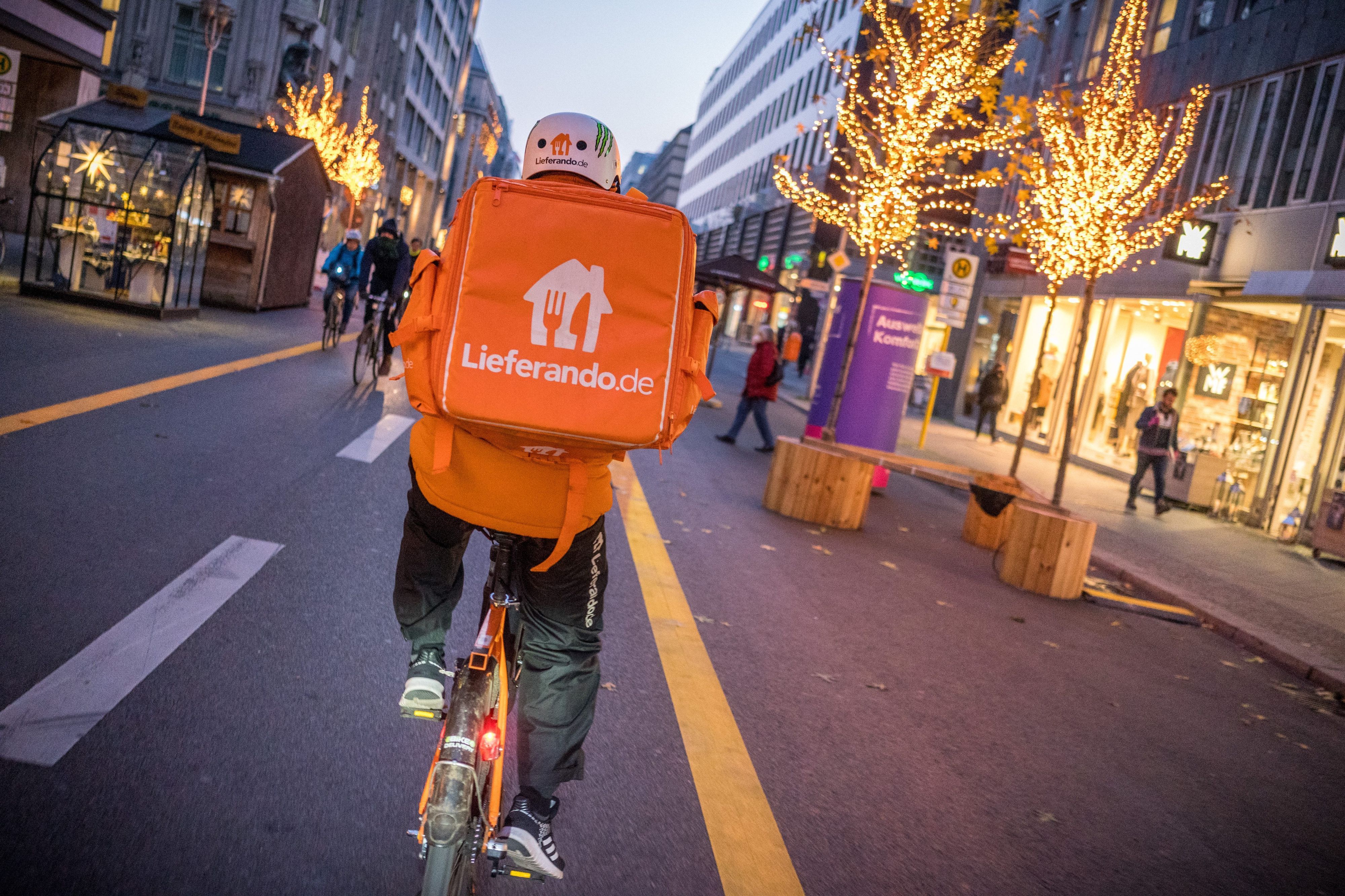 Ein Kurierfahrer des Restaurantlieferdiensts  Lieferando fährt mit seinem Fahrrad über die Friedrichstraße in Mitte. Er hat einen Lieferando-Rucksack auf dem Rücken. Auf der Straße ist Weihnachtsbeleuchtung.