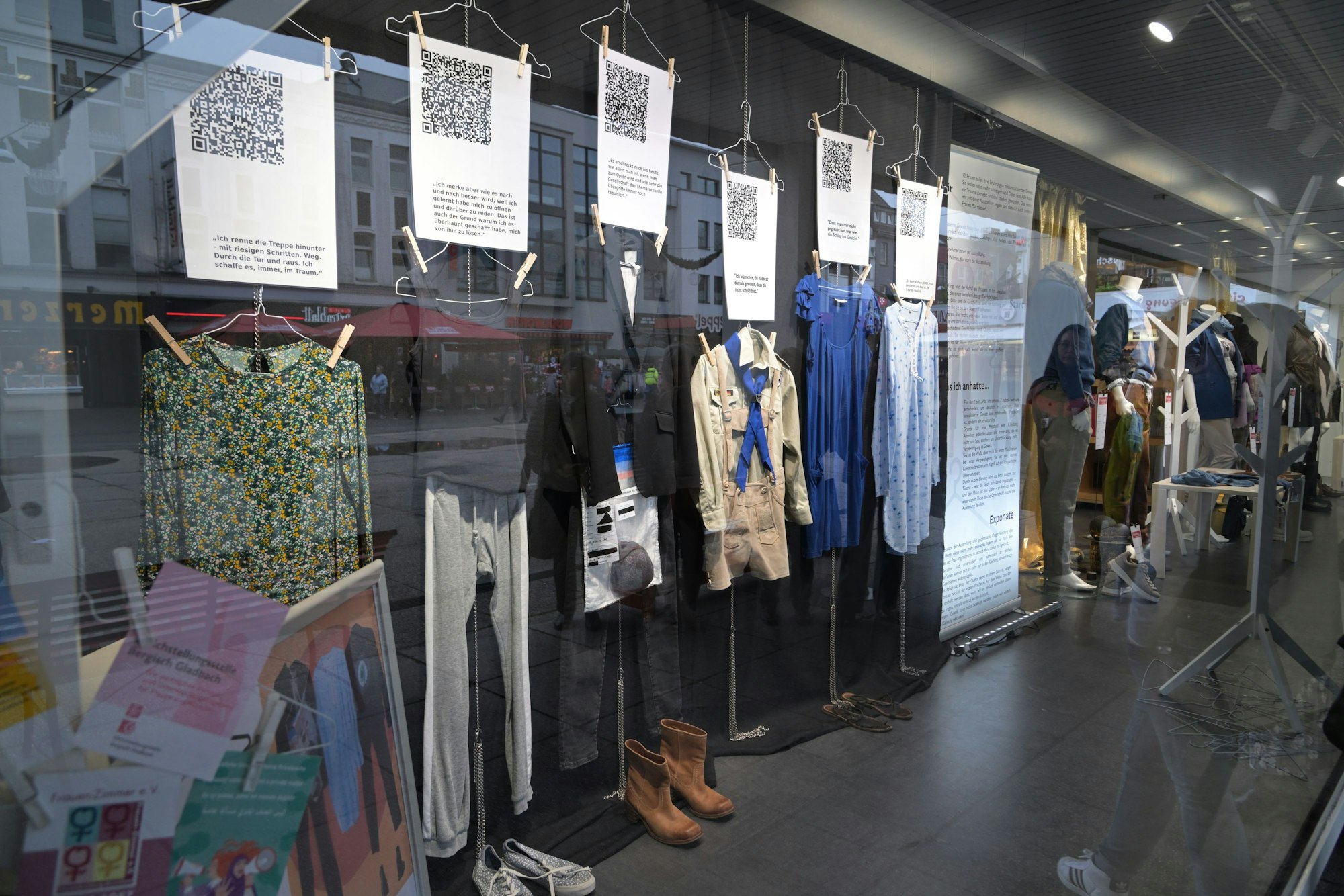 In einem Kleidungsgeschäft in Bergisch Gladbach sind an einer Wand verschiedene Kleidungsstücke ausgestellt. Über ihnen hängt jeweils ein großes Blatt Papier mit QR-Code.
