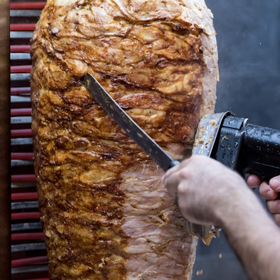 Jemand schneidet Fleisch mit einem elektrischen Gerät von einem Dönerspieß.