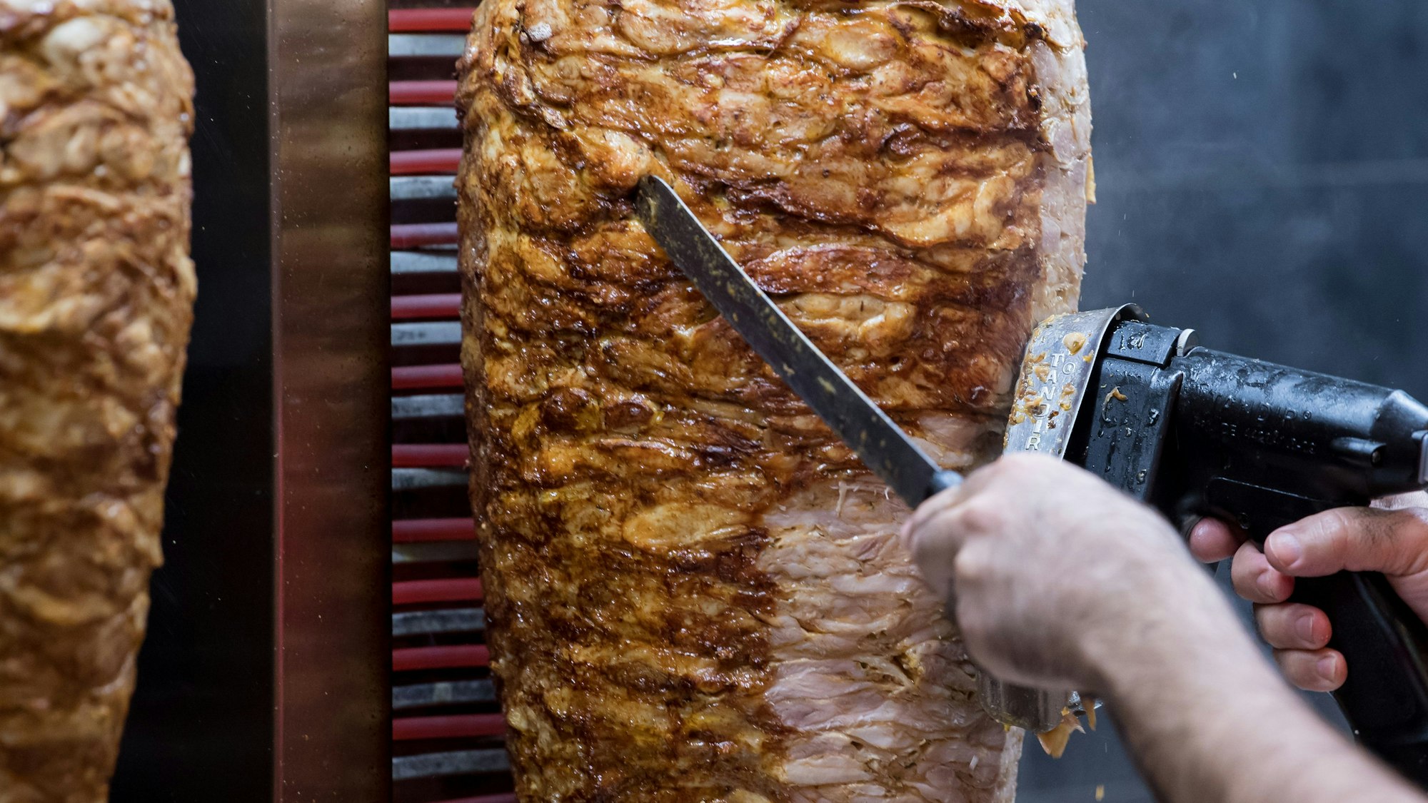 Jemand schneidet Fleisch mit einem elektrischen Gerät von einem Dönerspieß.