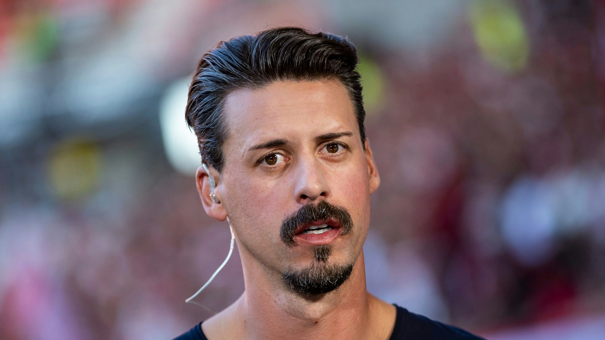 Der ehemalige deutsche Fußball-Nationalspieler Sandro Wagner trägt ein Headset und steht vor einem Spiel im Stadion. Er blickt in die Kamera.