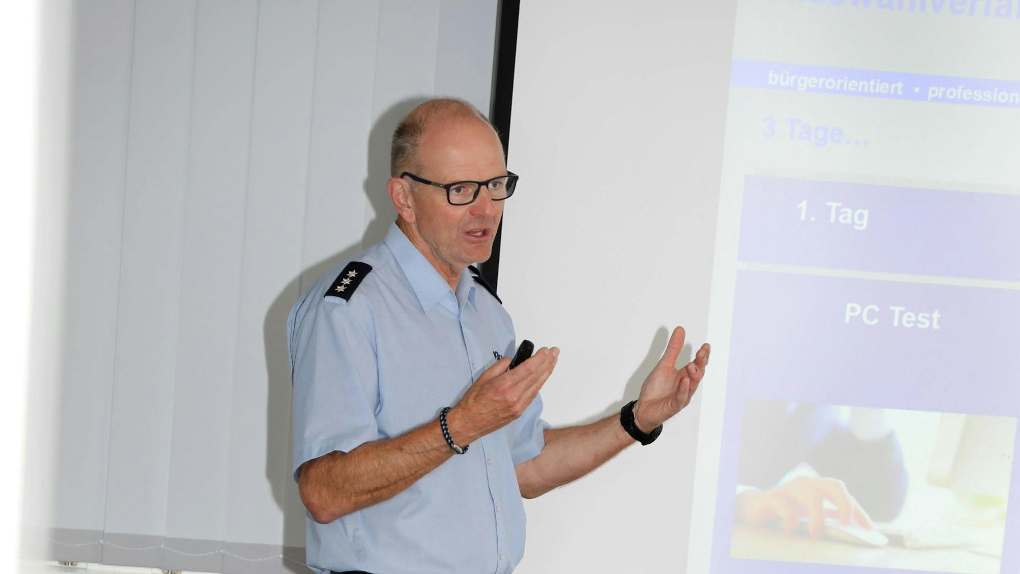 Peter Tilmans steht in Polizeihemd vor einer Leinwand mit einer Präsentation darauf und gestikuliert.