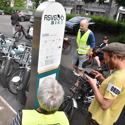 Menschen stehen um eine Stele der RSVG Bike-Vermietung herum. Dahinter sind E-Bikes aufgestellt.