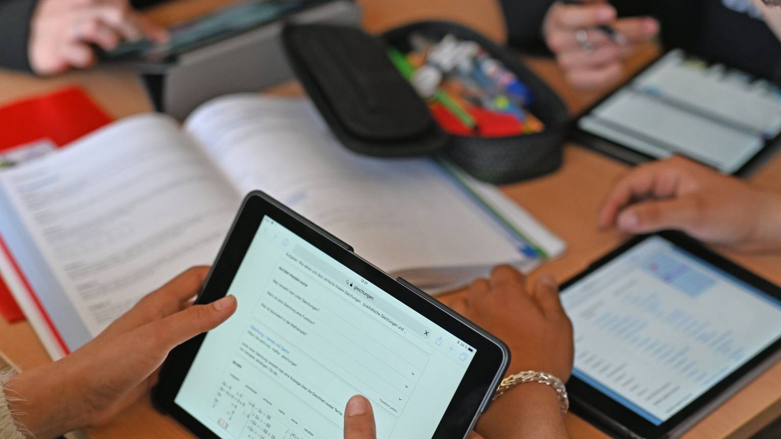 Auf einem Tisch liegen drei Tablets, auf denen getippt wird. Außerdem liegt ein Buch und ein Mäppchen mit Stiften auf dem Tisch.