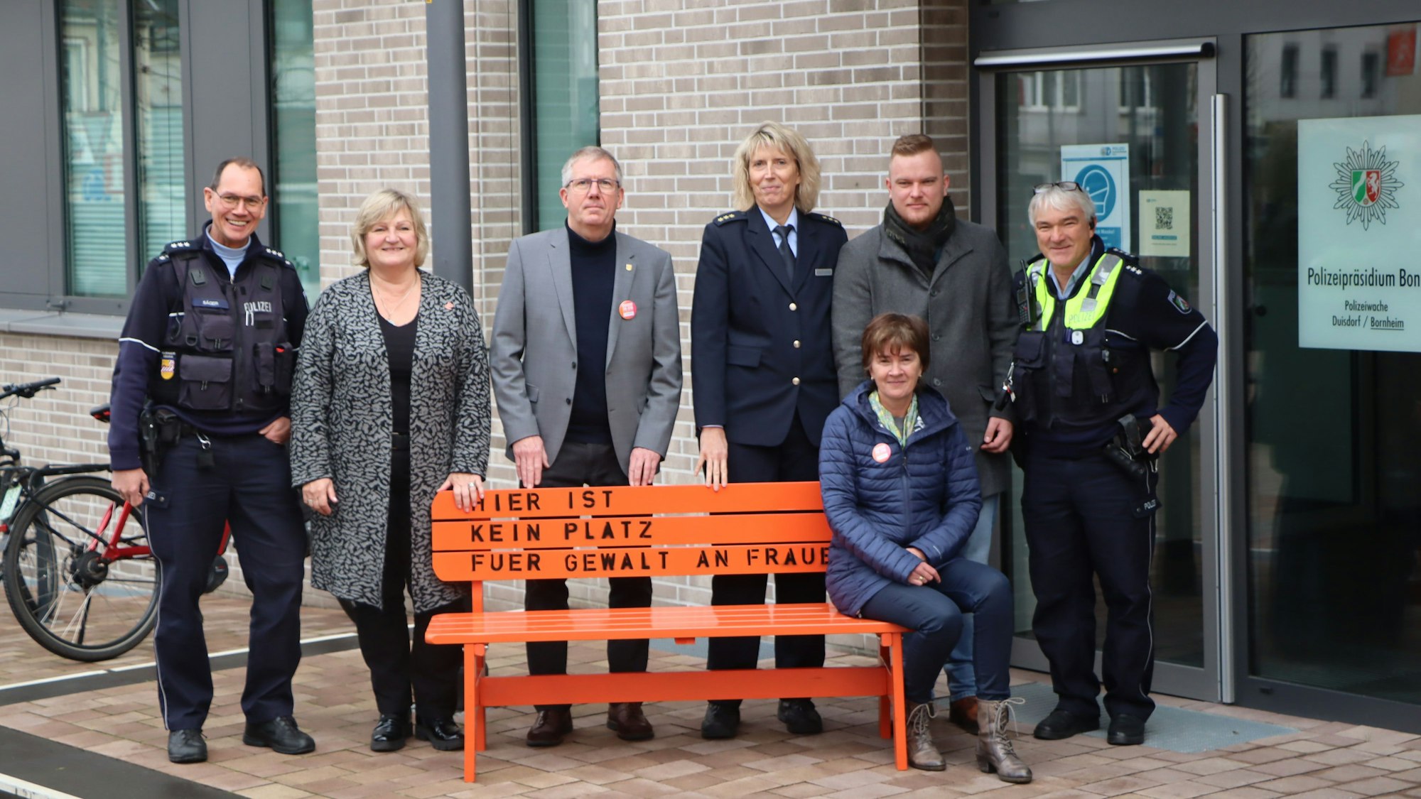 Vertreter der Polizei und der Stadt stehen am Peter-Fryns-Platz vor einer orangenen Bank mit der Aufschrift "Hier ist kein Platz für Gewalt an Frauen"