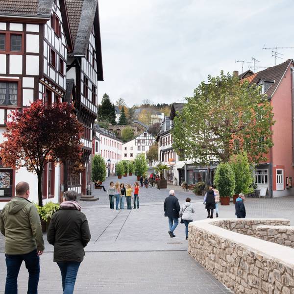 Das Bild zeigt den Marktplatz von Bad Münstereifel mit der Marktstraße im Hintergrund mit Blick von der Jesuitenkirche über die Heinz-Küpper-Brücke aus. Der Fotografie wurden Pflanzkübel und Bänke auf der Marktstraße hinzugefügt, die Autos wurden entfernt.