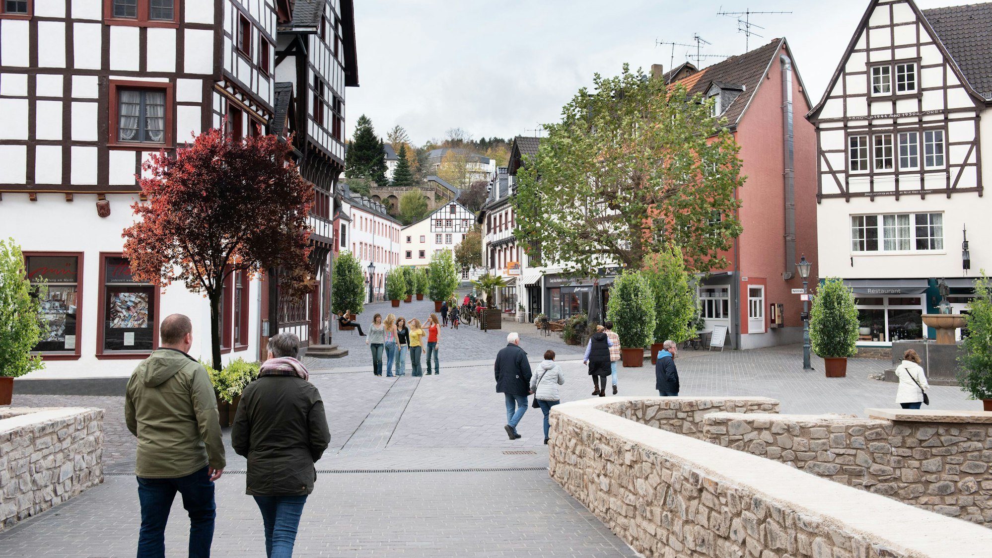 Das Bild zeigt den Marktplatz von Bad Münstereifel mit der Marktstraße im Hintergrund mit Blick von der Jesuitenkirche über die Heinz-Küpper-Brücke aus. Der Fotografie wurden Pflanzkübel und Bänke auf der Marktstraße hinzugefügt, die Autos wurden entfernt.