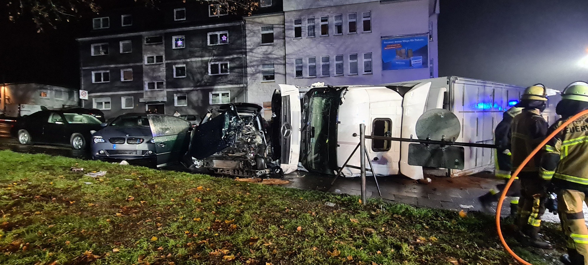Zerstörte Autos stehen auf einer Straße vor Wohnhäusern, ein großer weißer Lkw liegt auf der Seite, am rechten Bildrand stehen Feuerwehrleute.