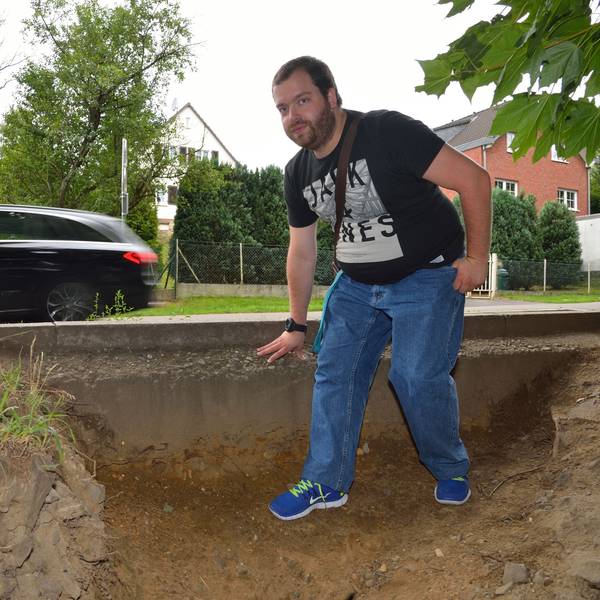 Ein junger Mann im schwarzen T-Shirt steht auf dem Trampelpfad am Rösrather Bahnhof. Im Hintergrund fährt ein Auto vorbei.