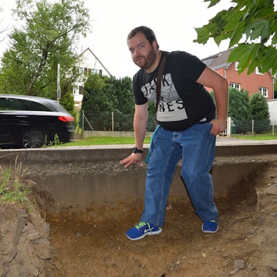 Ein junger Mann im schwarzen T-Shirt steht auf dem Trampelpfad am Rösrather Bahnhof. Im Hintergrund fährt ein Auto vorbei.