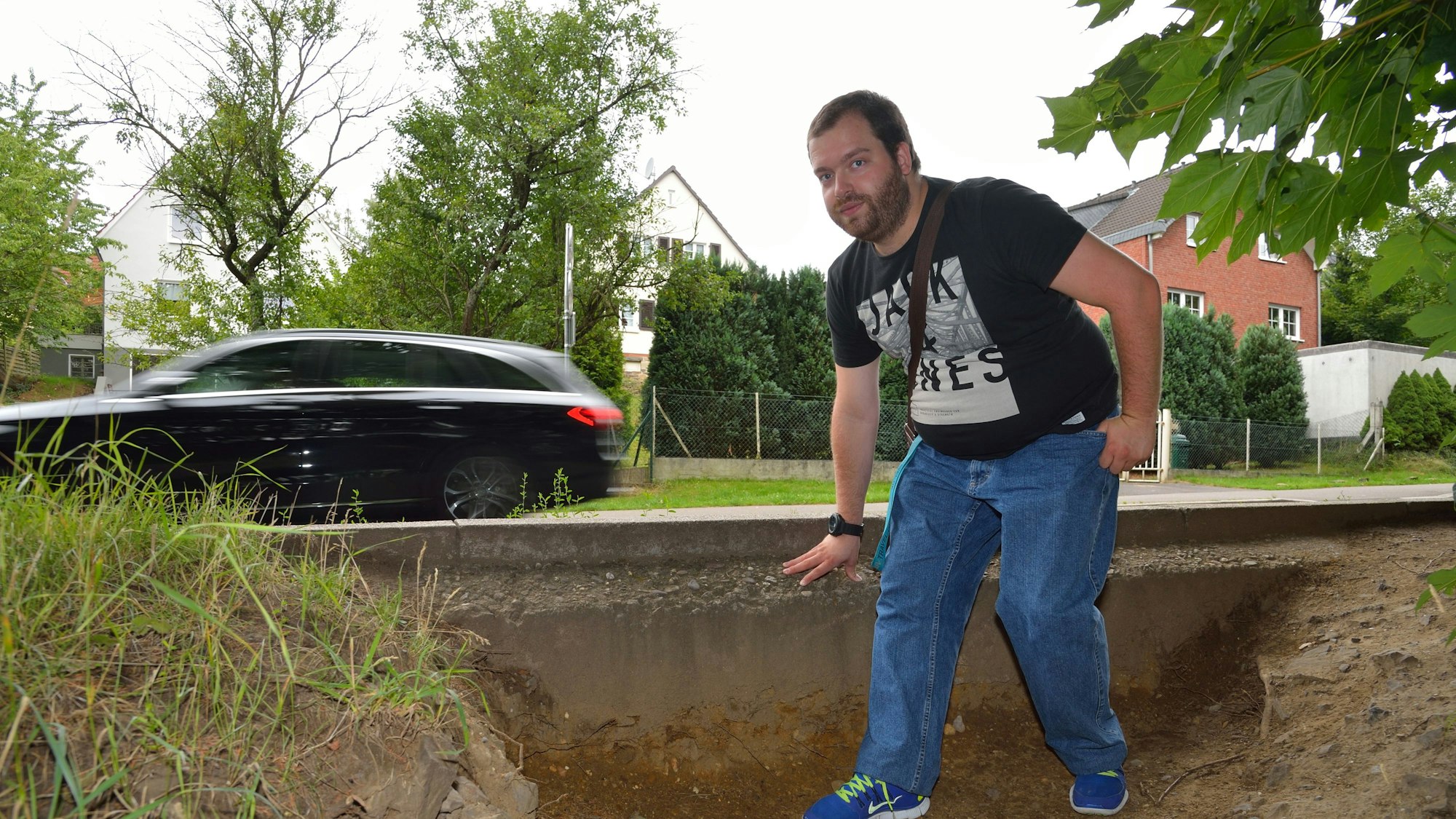 Ein junger Mann im schwarzen T-Shirt steht auf dem Trampelpfad am Rösrather Bahnhof. Im Hintergrund fährt ein Auto vorbei.