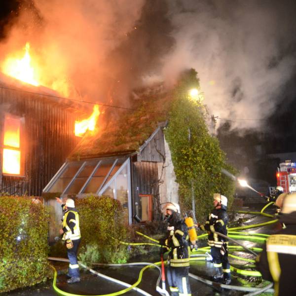 Feuerwehrleute stehen vor einem Haus mit Holzverkleidung, das in Flammen steht. Das Feuer kommt aus einem Fenster und dem Dach. In einem anderen Fenster ist deutlich zusehen, wie es im Inneren des Hauses brennt.