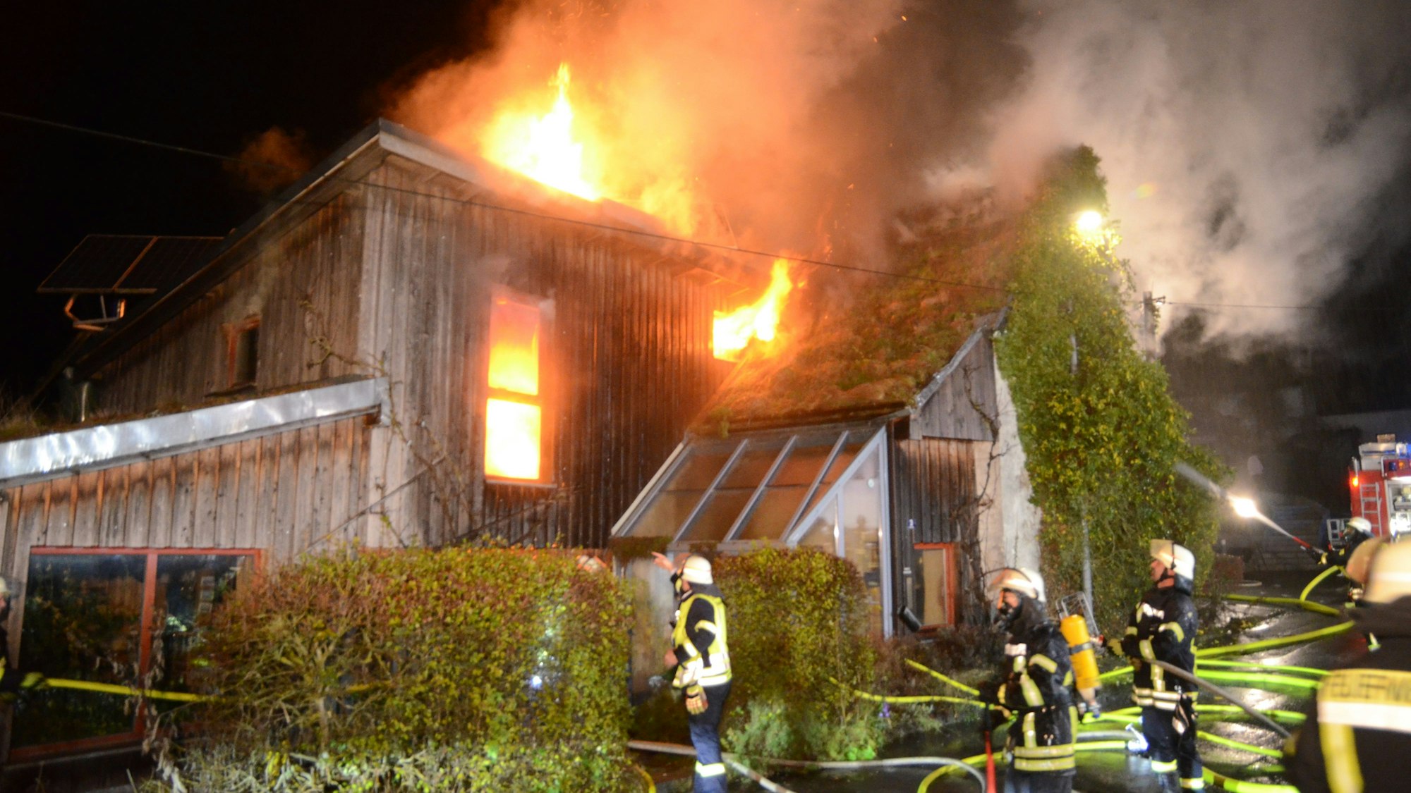 Feuerwehrleute stehen vor einem Haus mit Holzverkleidung, das in Flammen steht. Das Feuer kommt aus einem Fenster und dem Dach. In einem anderen Fenster ist deutlich zusehen, wie es im Inneren des Hauses brennt.