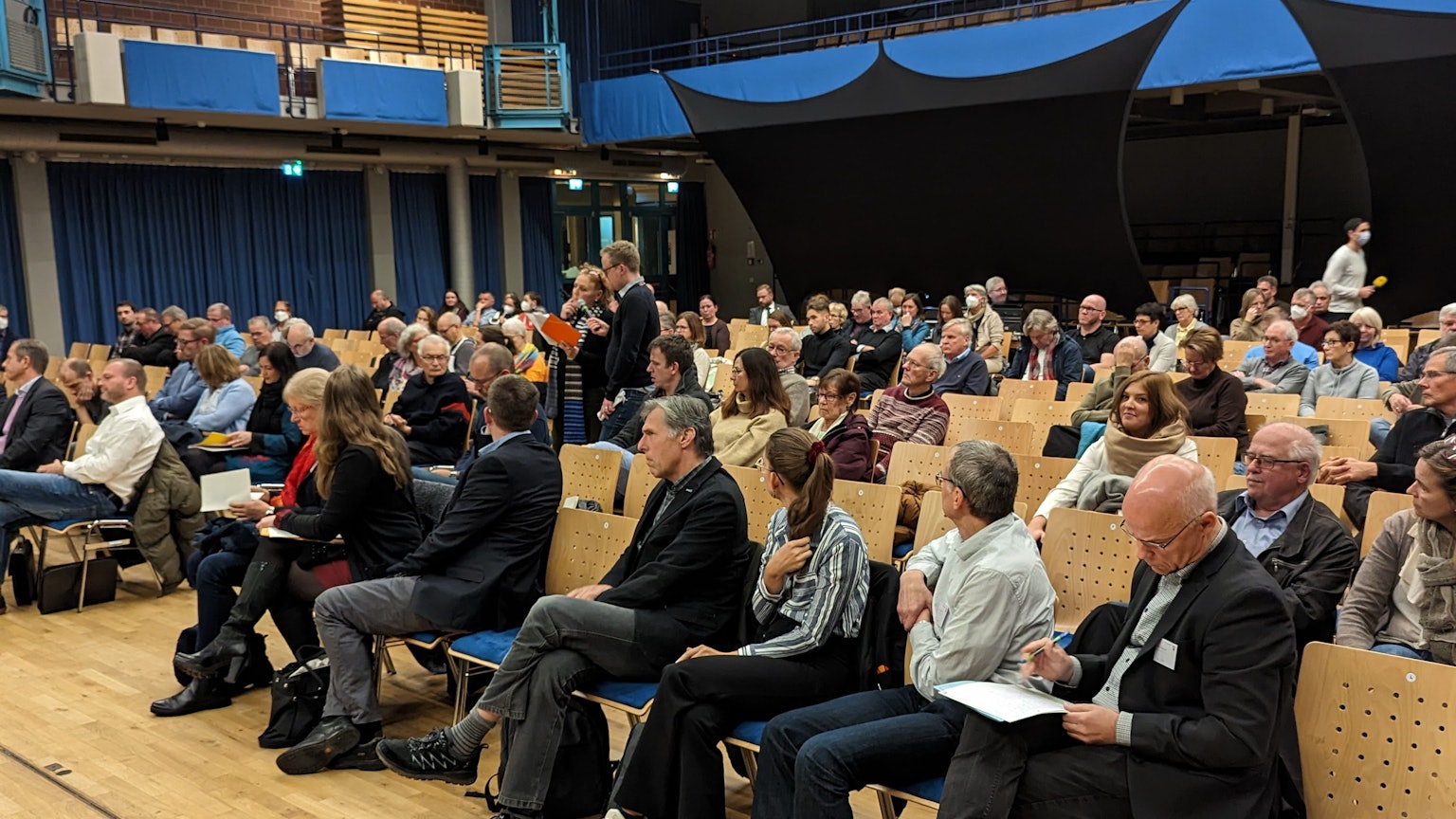 In einer Halle stehen mehrere Stuhlreihen. Viele Personen sitzen auf den Stühlen.