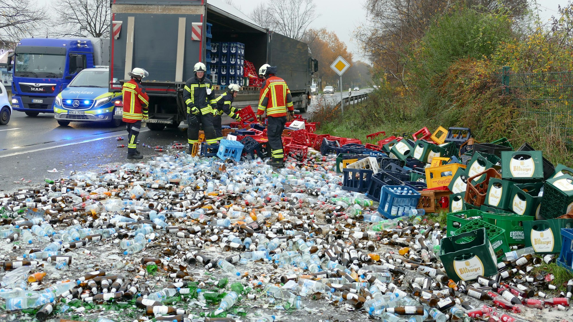 Ein Lkw steht am Straßenrand, eine Seite ist geöffnet. Davor liegen unzählig viele kaputte Getränkeflaschen und -Kisten. Drei Feuerwehrmänner räumen auf.