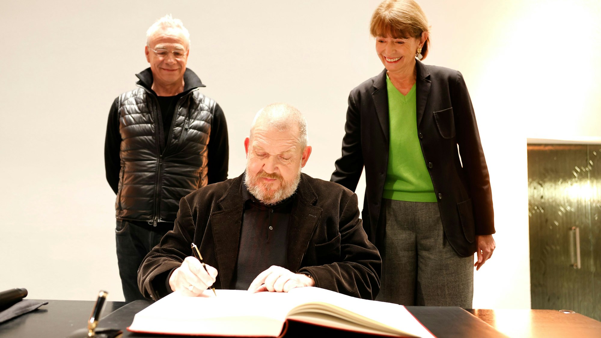 Dietmar Bär trägt sich ins Goldene Buch ein, Klaus J. Behrendt und Henriette Reker schauen zu.