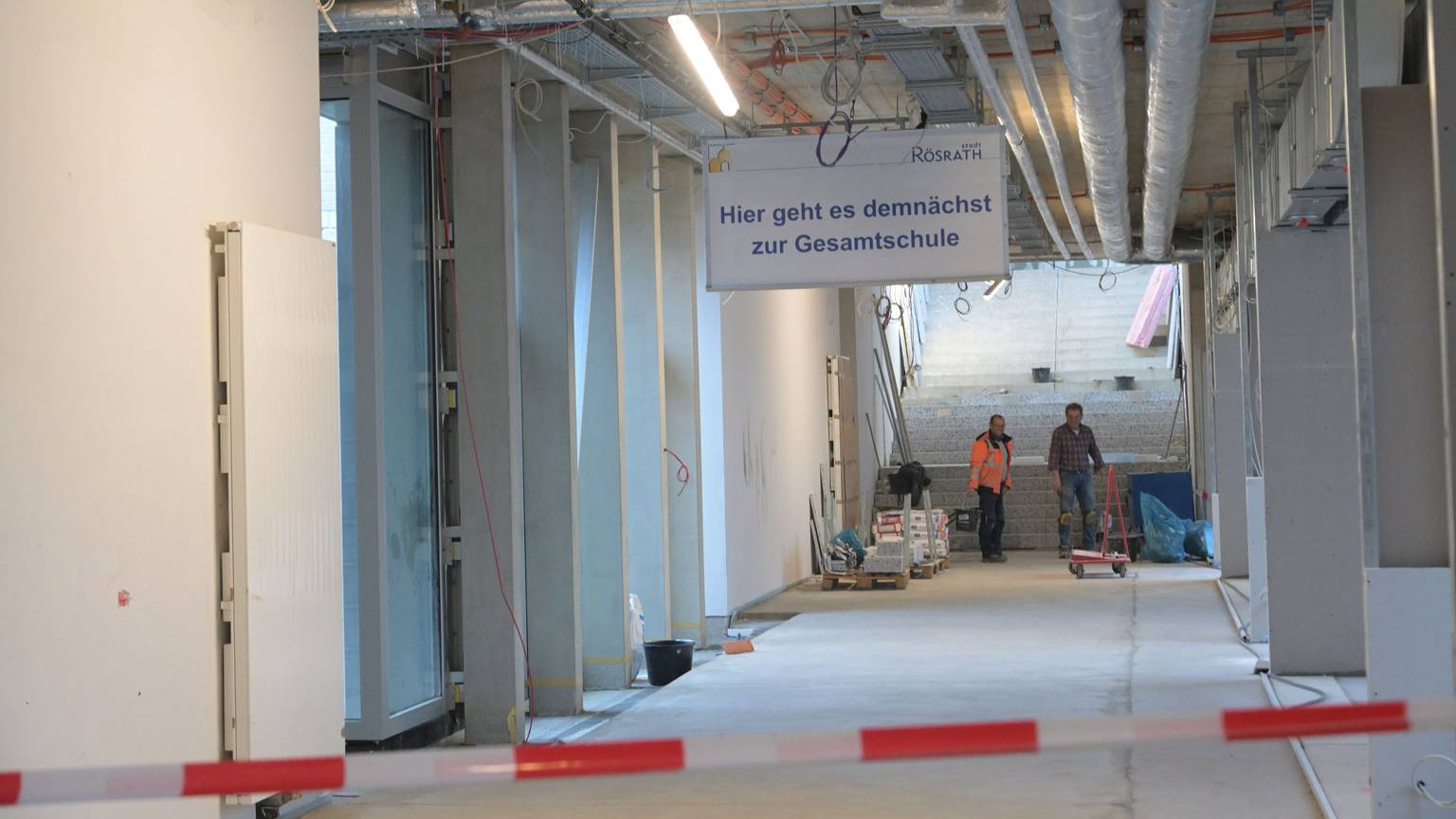 Ein noch im Bau befindlicher Flur im Rösrather Schulzentrun, an dessen Ende zwei Arbeiter stehen. An der Decke hängt ein Schild mit der Aufschrift "Hier geht es demnächst zur Gesamtschule".