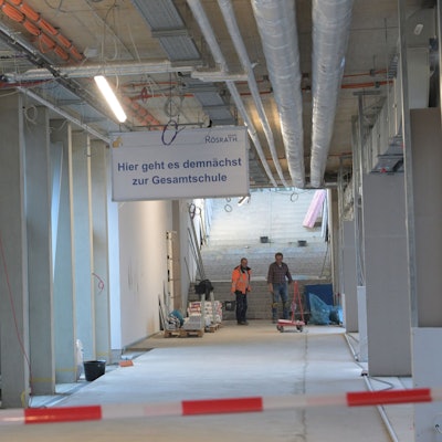Ein noch im Bau befindlicher Flur im Rösrather Schulzentrun, an dessen Ende zwei Arbeiter stehen. An der Decke hängt ein Schild mit der Aufschrift "Hier geht es demnächst zur Gesamtschule".