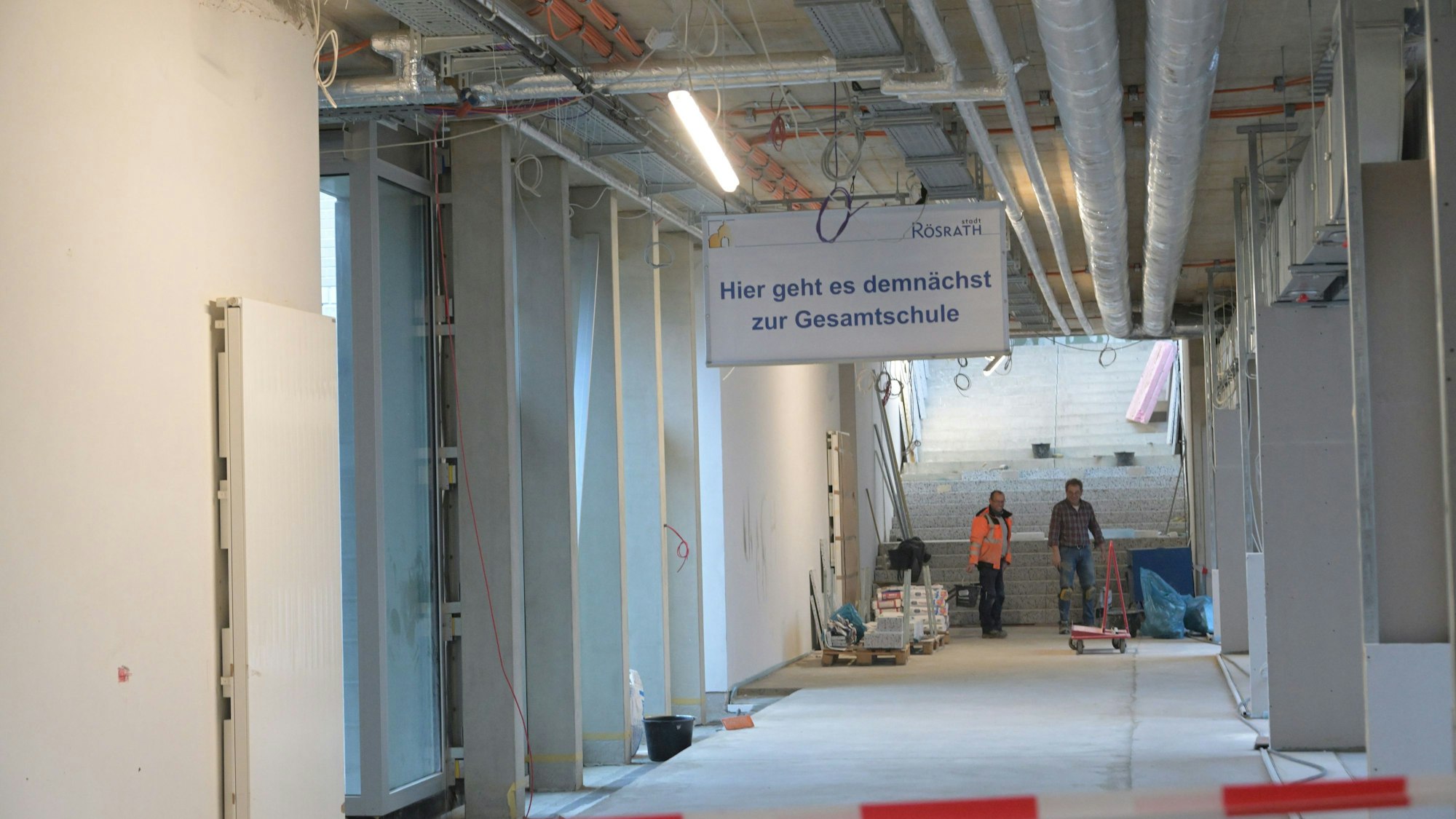 Ein noch im Bau befindlicher Flur im Rösrather Schulzentrun, an dessen Ende zwei Arbeiter stehen. An der Decke hängt ein Schild mit der Aufschrift "Hier geht es demnächst zur Gesamtschule".
