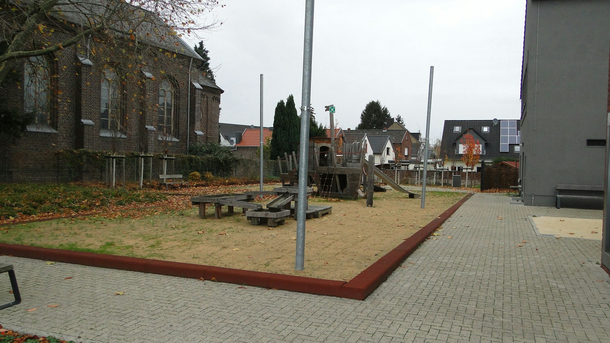Spielgerüste stehen auf einem Hof, im Hintergrund sind Häuser, links daneben ein Schulgebäude.