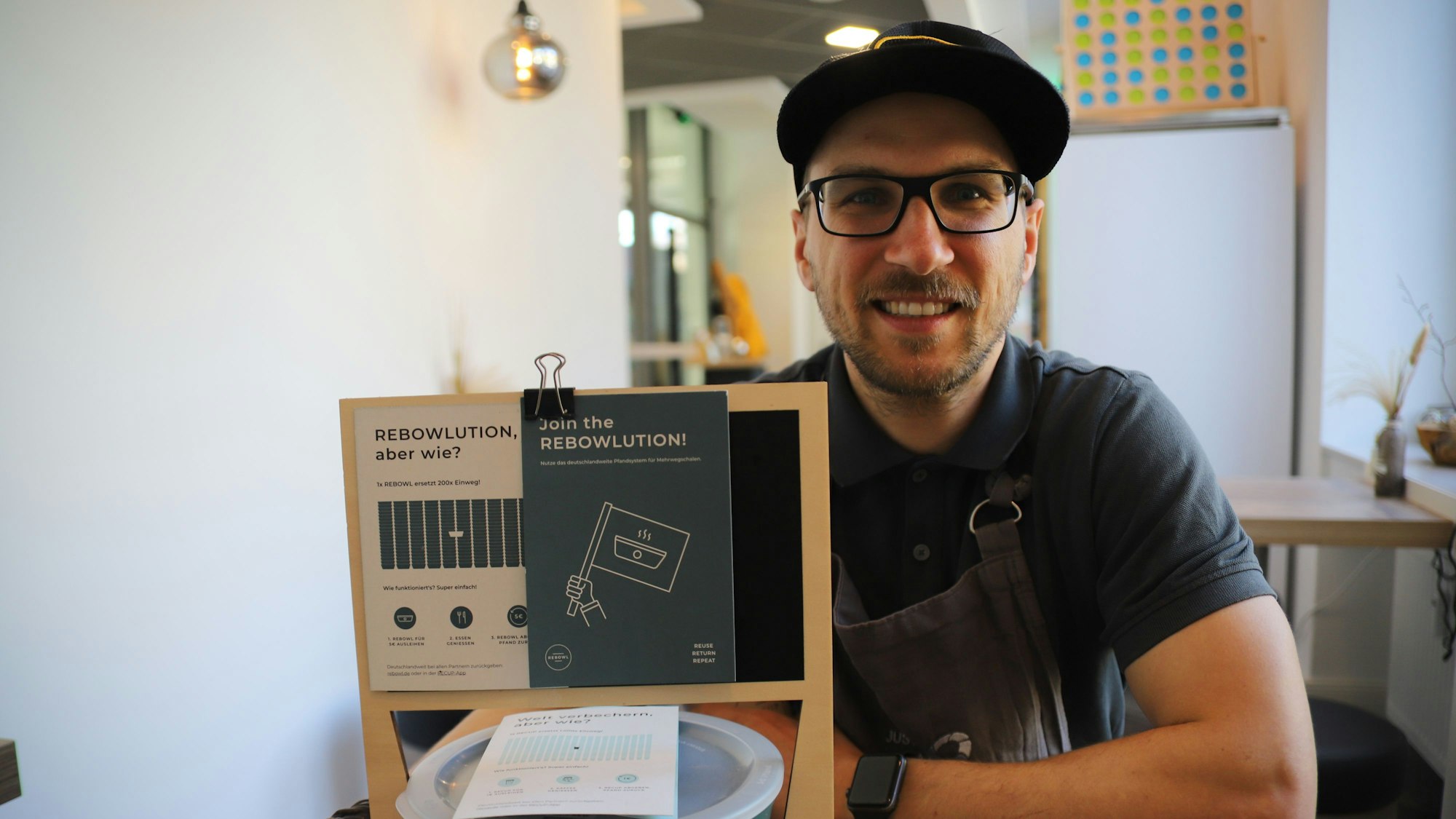 Der Besitzer des Hennefer Lokals „Just Be Healthy“ sitzt, vor ihm stehen Informationsblätter über das Einweg-Geschirr.