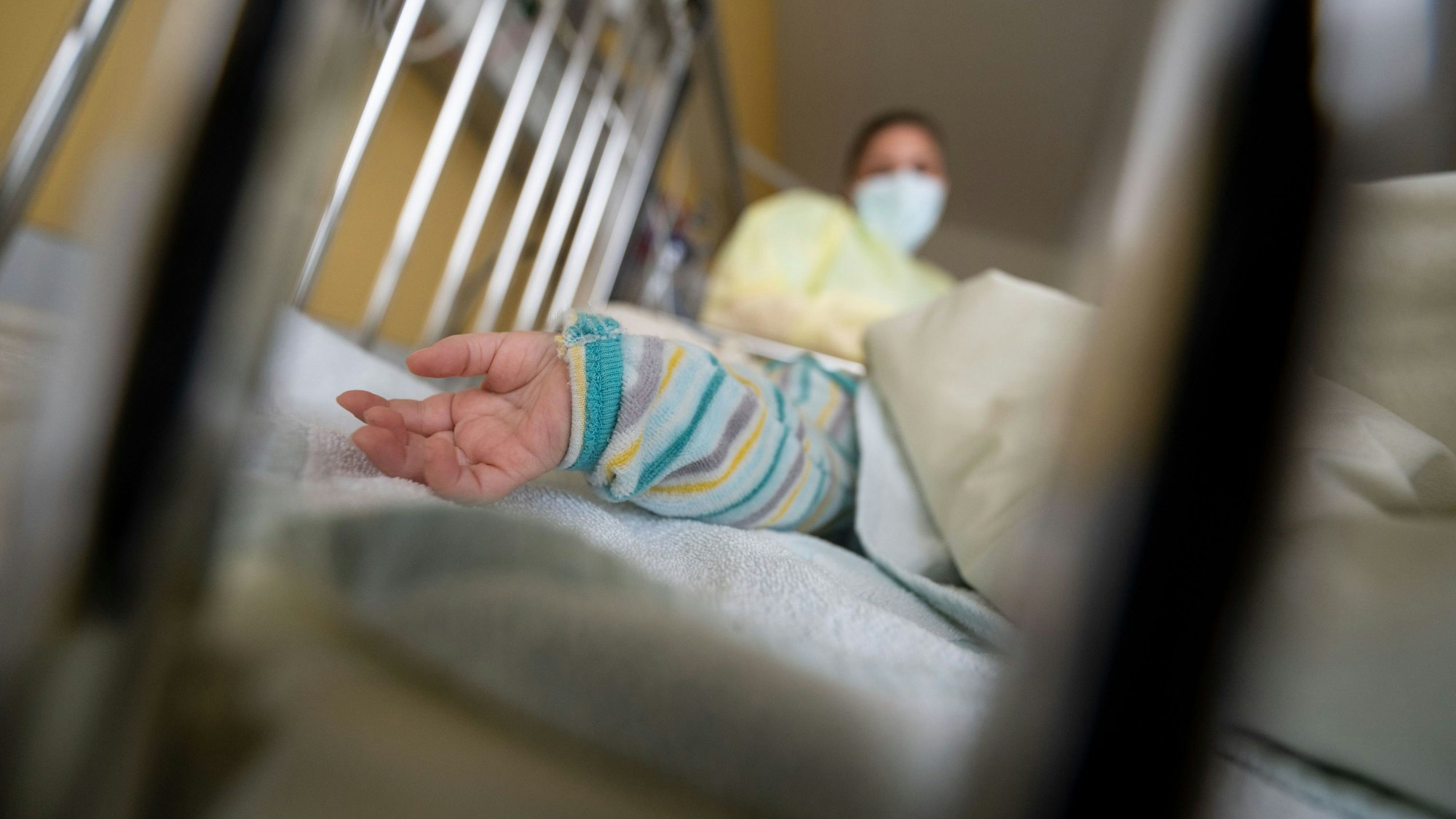 In einem Gitterbett ist eine Kinderhand zu sehen. Im Hintergrund steht eine Frau mit Mundschutz und Kittel.