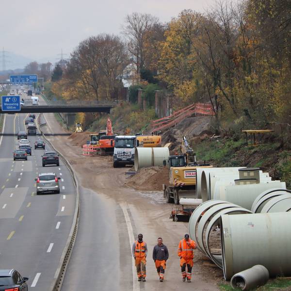 Auf einer Autobahn fahren Fahrzeuge auf drei verengten Fahrstreifen. Am rechten Fahrbahnrand laufen drei Baustellenarbeiter mit Warnkleidung über den abgesperrten Standstreifen. Im Hintergrund stehen Bagger.