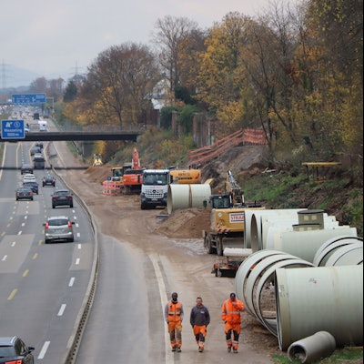 Auf einer Autobahn fahren Fahrzeuge auf drei verengten Fahrstreifen. Am rechten Fahrbahnrand laufen drei Baustellenarbeiter mit Warnkleidung über den abgesperrten Standstreifen. Im Hintergrund stehen Bagger.