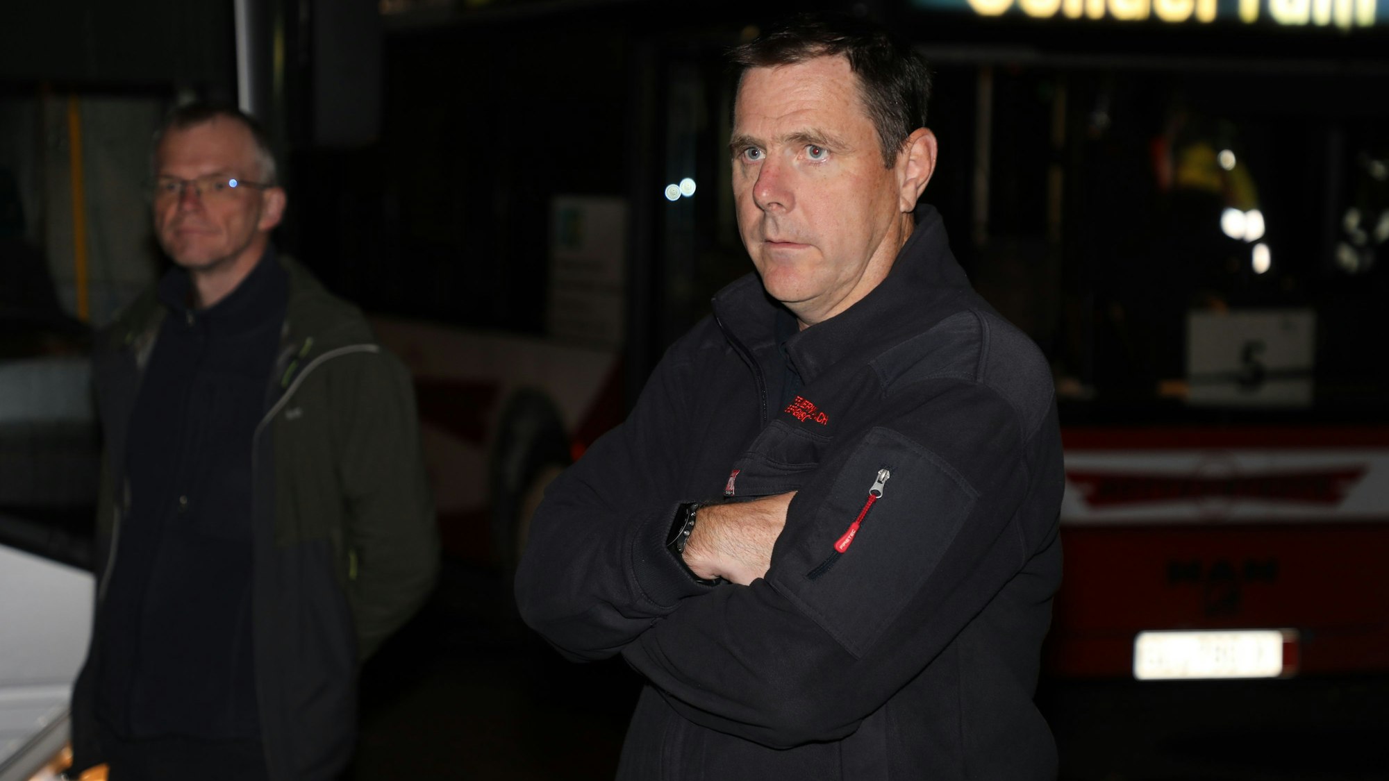 Feuerwehrchef Jörg Köhler steht im Dunkeln mit verschränkten Armen vor einem Bus, dessen Anzeige „Sonderfahrt“ anzeigt. Im Hintergrund steht Bürgermeister Frank Stein.