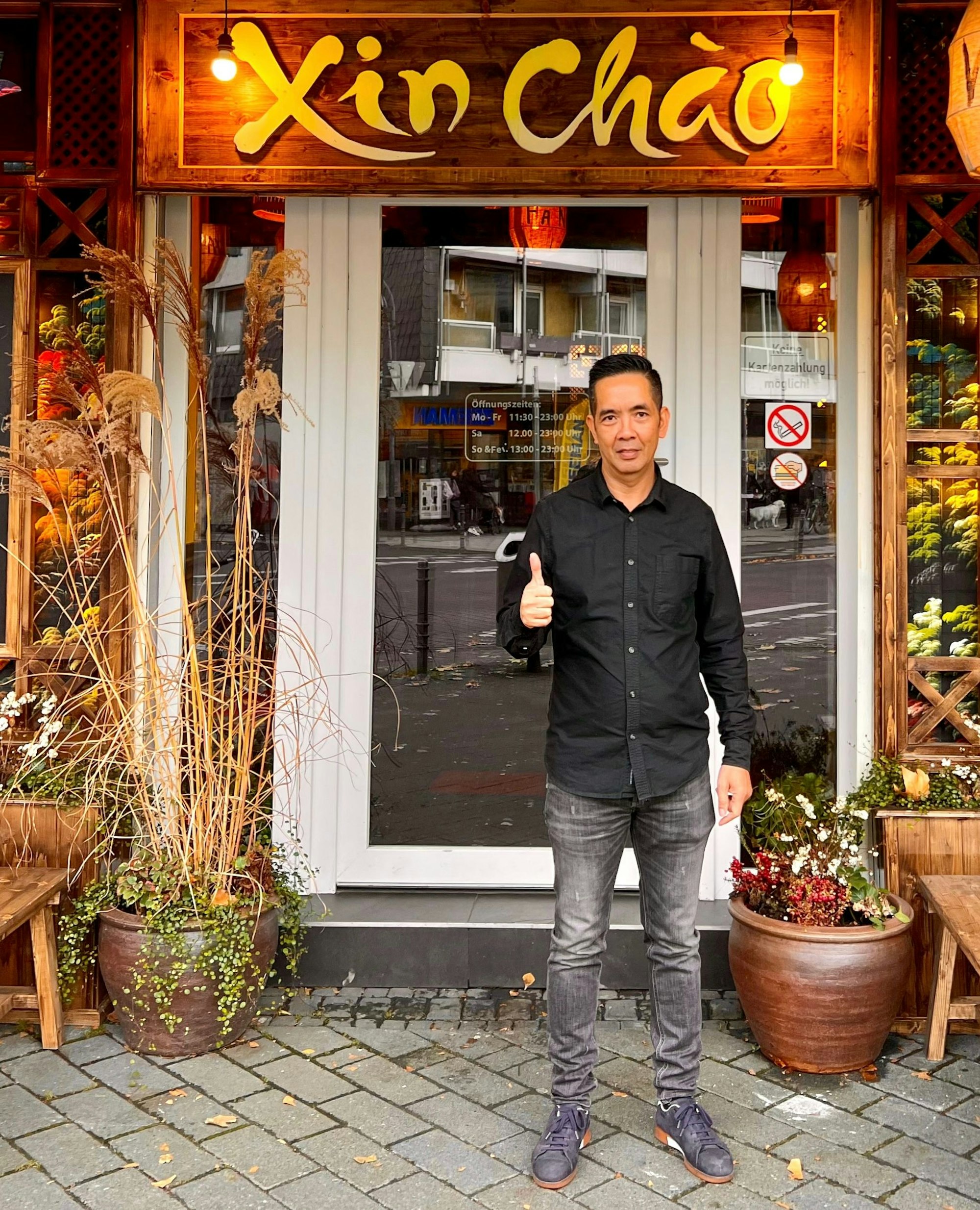 Ein Mann mit dunklem Haaren steht vor der Tür zu einem Restaurant. Er macht die Geste „Daumen hoch".