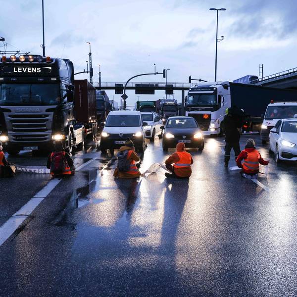 Klimaaktivisten sitzen mit dem Rücken zur Kamera auf einer mehrspurigen Kreuzung. Vor ihnen stehen Autos und LKW.