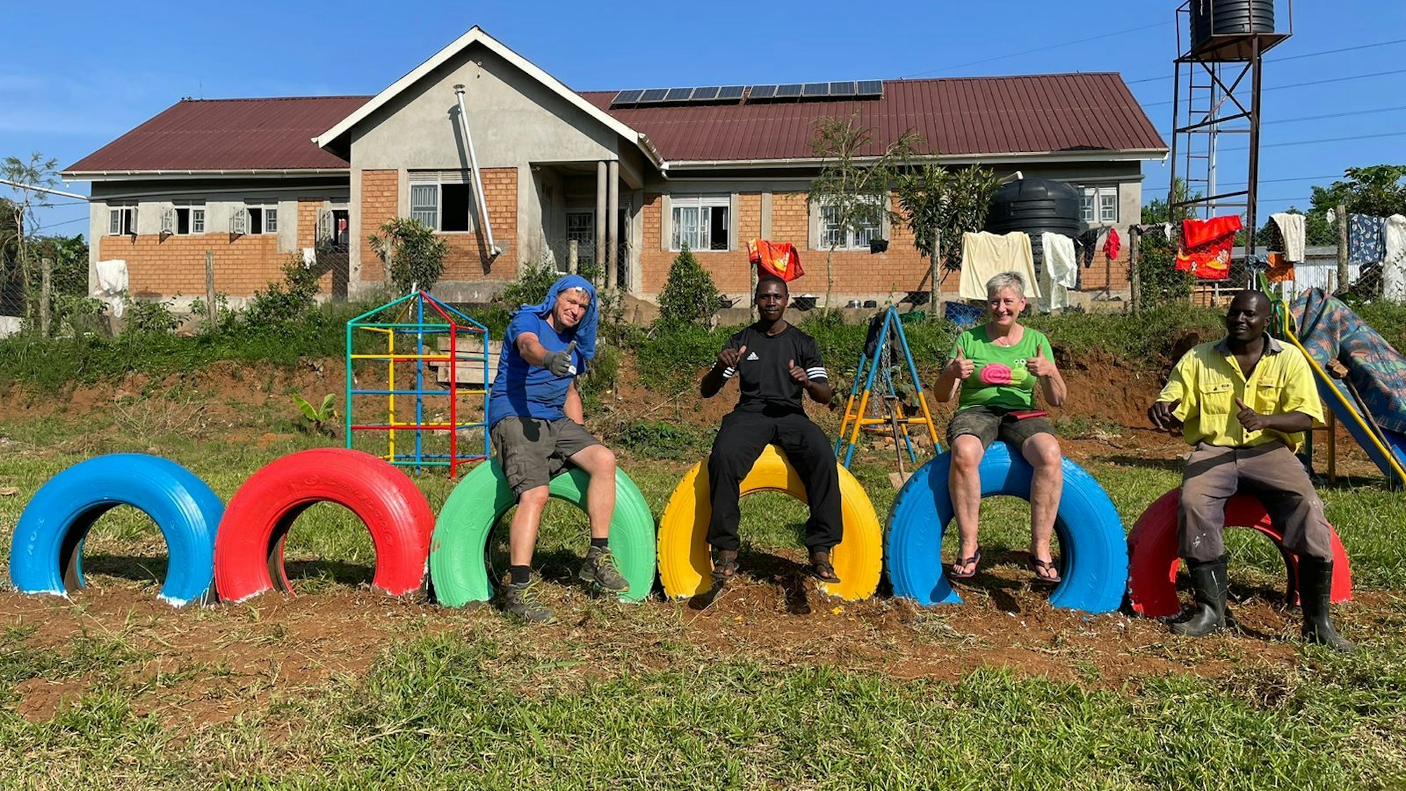 Vier Personen sitzen auf bunten Autoreifen. Im Hintergrund sind Spielgeräte für Kinder zu sehen und ein Haus.