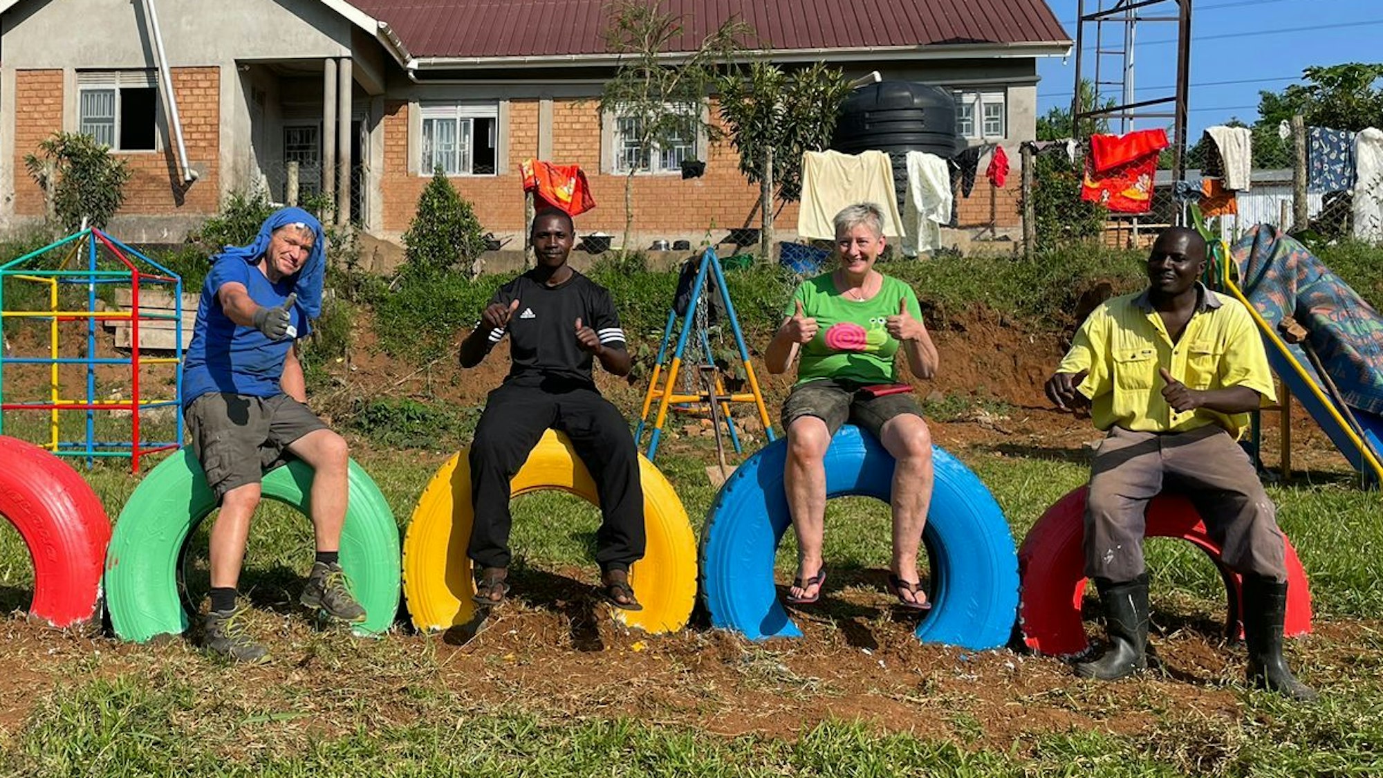 Vier Menschen sitzen auf bunt bemalten Lkw-Reifen vor einem Gebäude auf einem Spielplatz.