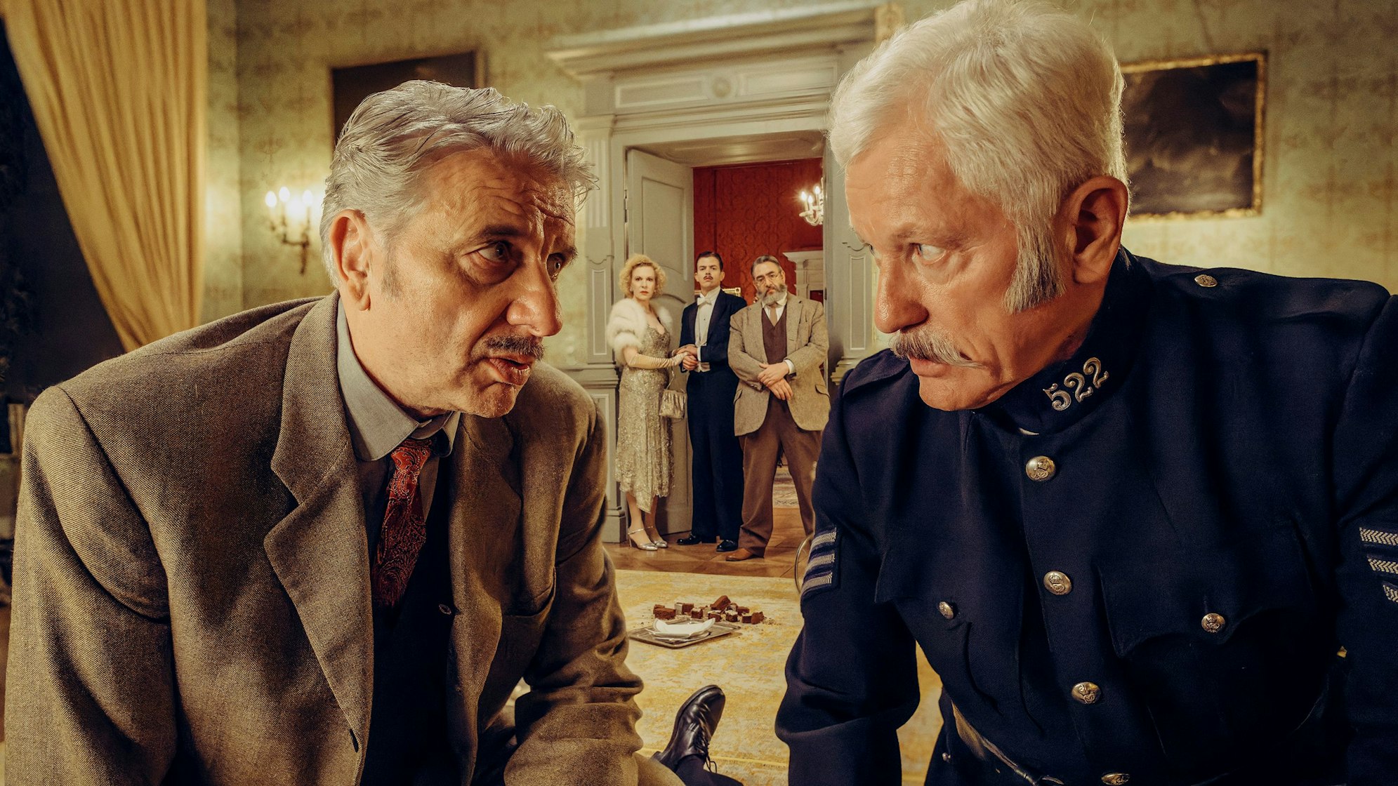 Chief Inspector Francis Lightmyer (Udo Wachtveitl, links vorne) und Constable Ivor Partridge (Miroslav Nemec, rechts vorne) untersuchen Arthur im Kaminzimmer. Lady Bantam (Sunnyi Melles), Charles (Ferdinand Hofer) und Dr. Mallard (Alexander Hörbe, rechts hinten) beobachten sie.