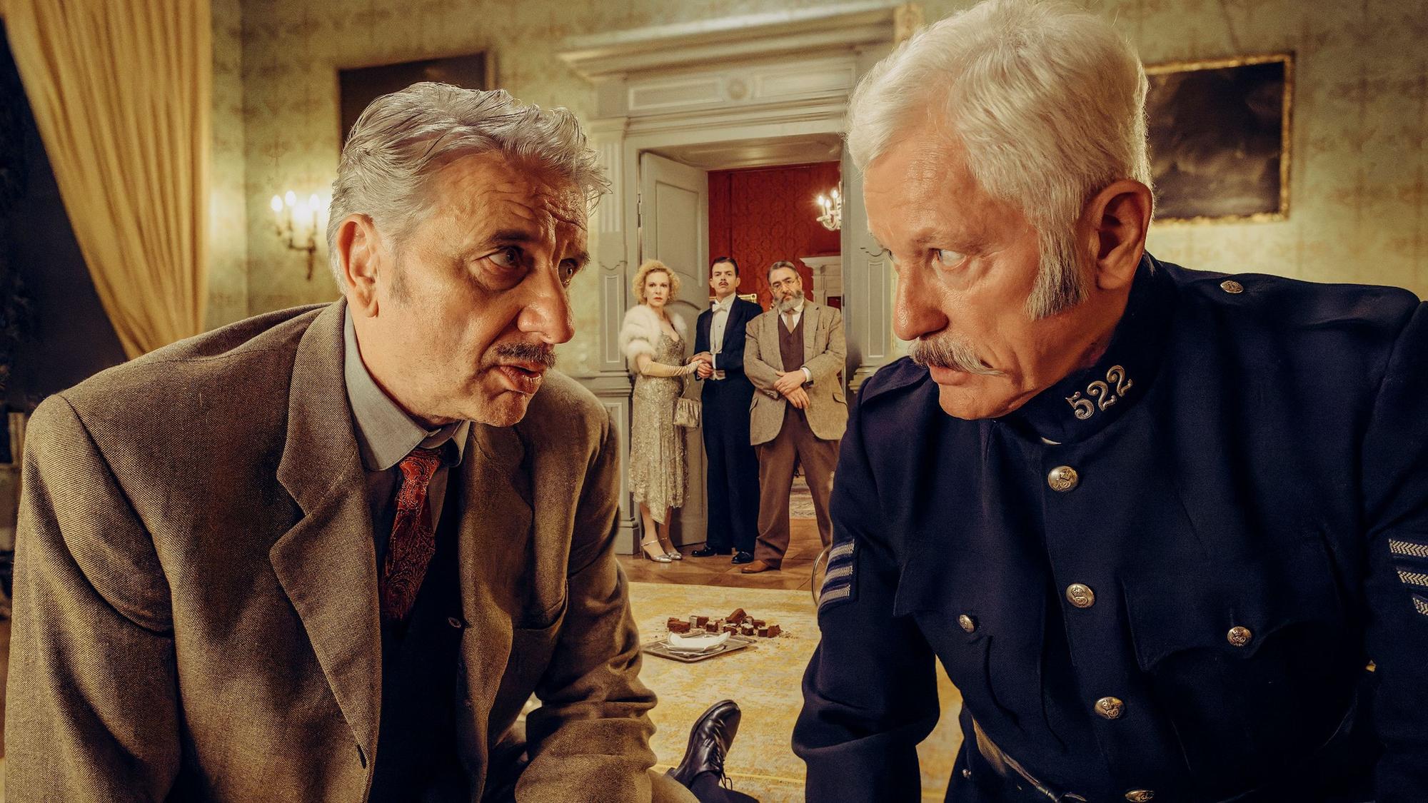 Chief Inspector Francis Lightmyer (Udo Wachtveitl, links vorne) und Constable Ivor Partridge (Miroslav Nemec, rechts vorne) untersuchen Arthur im Kaminzimmer. Lady Bantam (Sunnyi Melles), Charles (Ferdinand Hofer) und Dr. Mallard (Alexander Hörbe, rechts hinten) beobachten sie.
