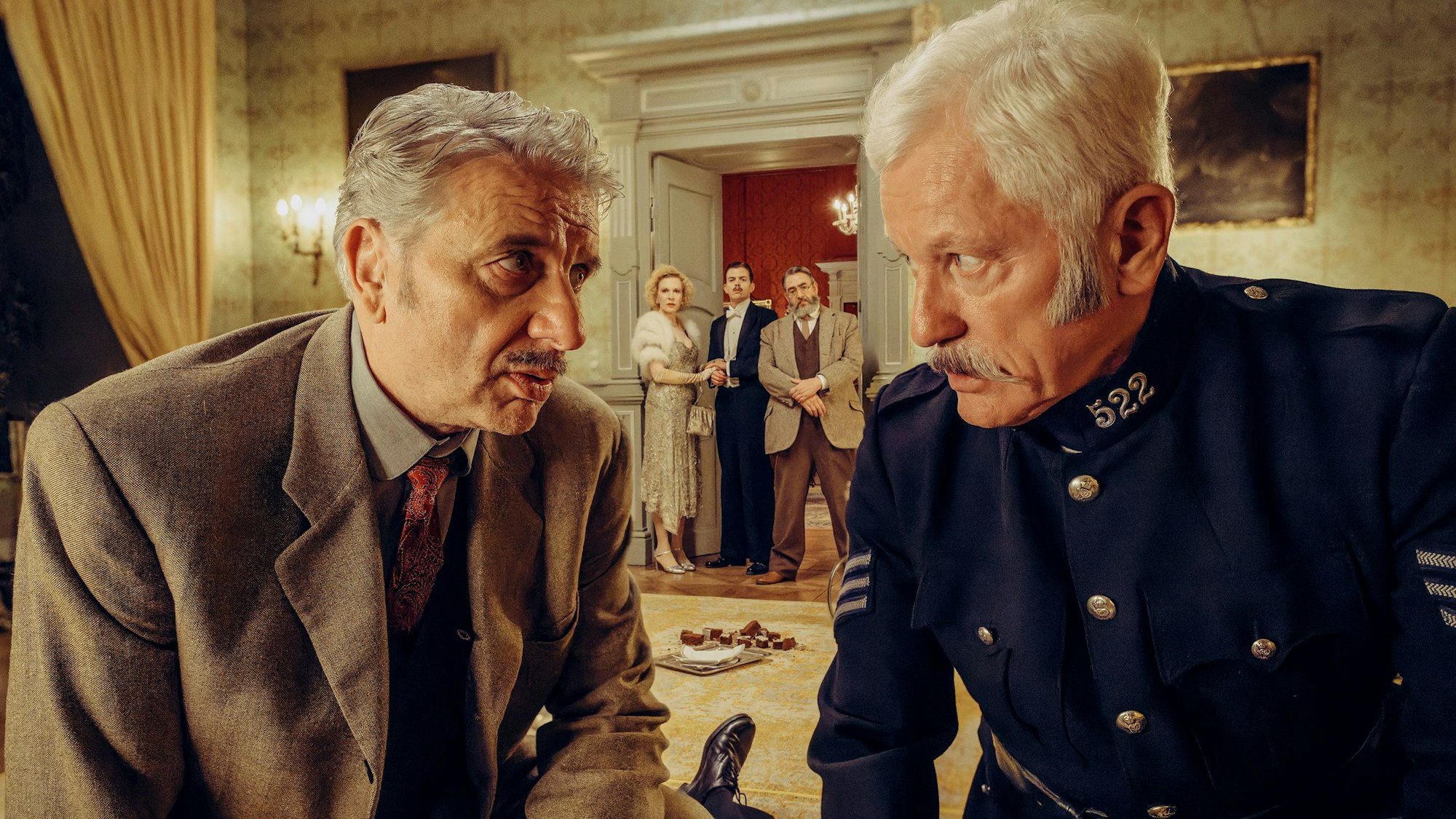 Chief Inspector Francis Lightmyer (Udo Wachtveitl, links vorne) und Constable Ivor Partridge (Miroslav Nemec, rechts vorne) untersuchen Arthur im Kaminzimmer. Lady Bantam (Sunnyi Melles), Charles (Ferdinand Hofer) und Dr. Mallard (Alexander Hörbe, rechts hinten) beobachten sie.