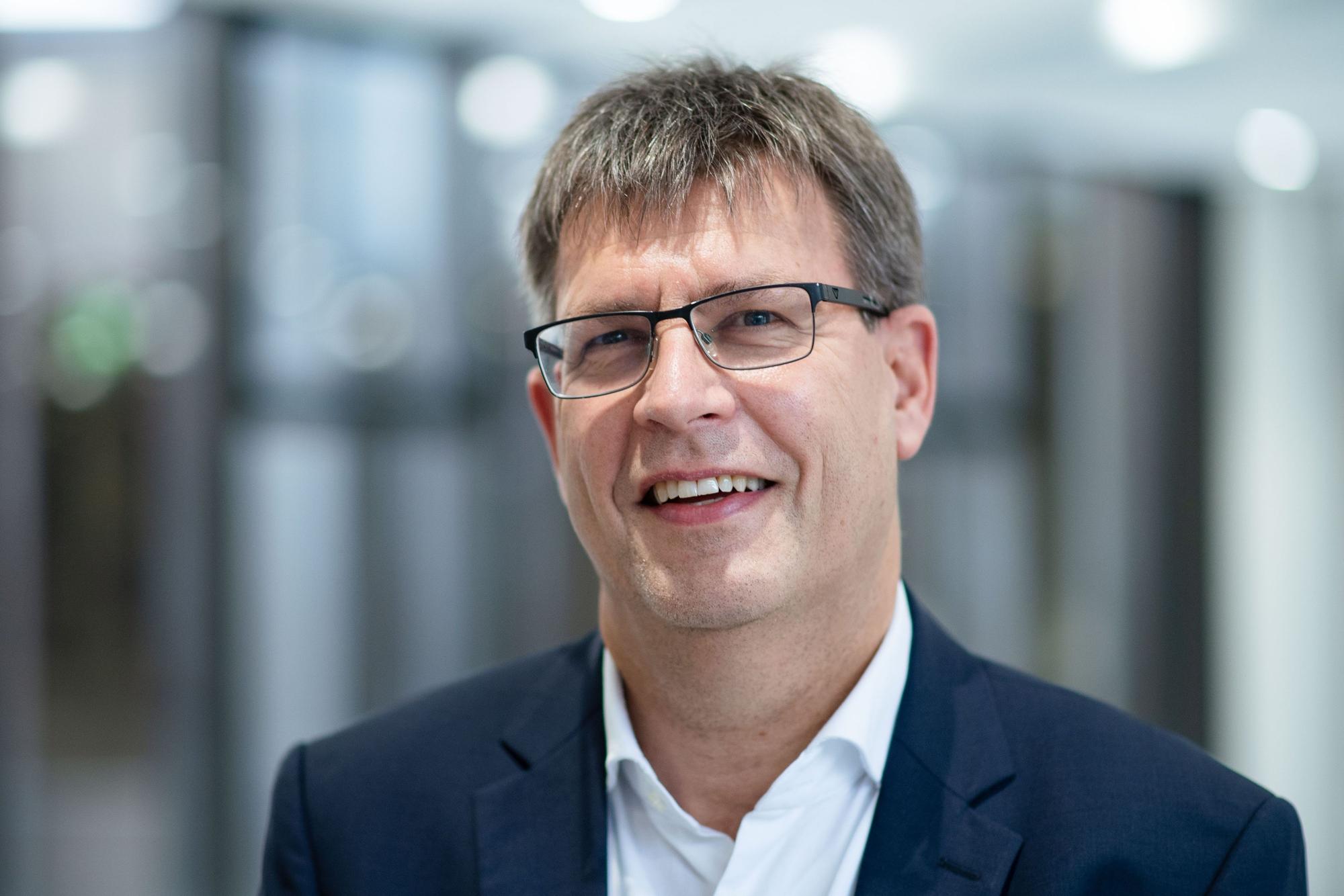 Thomas Weikert mit weißem Hemd und blauem Sakko, trägt eine Brille und Kurzhaarfrisur, lächelt in die Kamera.