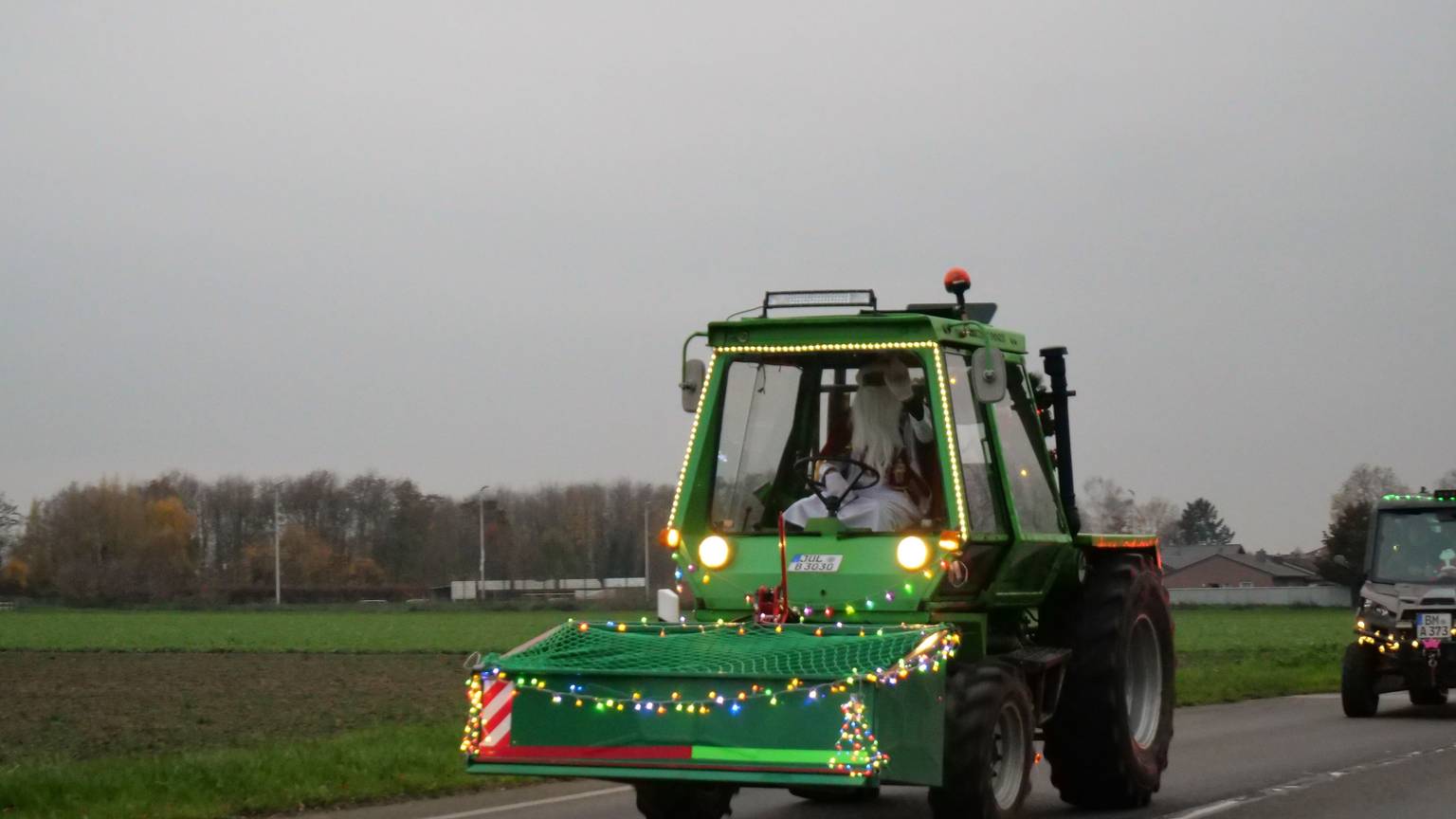 Ein geschmückter, grüner Traktor fährt über eine Straße. Am Steuer sitzt ein Mann, der als Weihnachtsmann verkleidet ist.