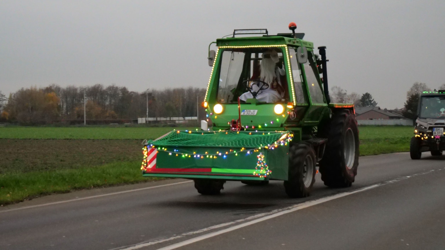 Ein geschmückter, grüner Traktor fährt über eine Straße. Am Steuer sitzt ein Mann, der als Weihnachtsmann verkleidet ist.