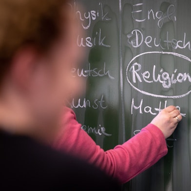 Eine Lehrerin schreibt mit Kreide einen Stundenplan mit Religionsunterricht an eine Tafel.