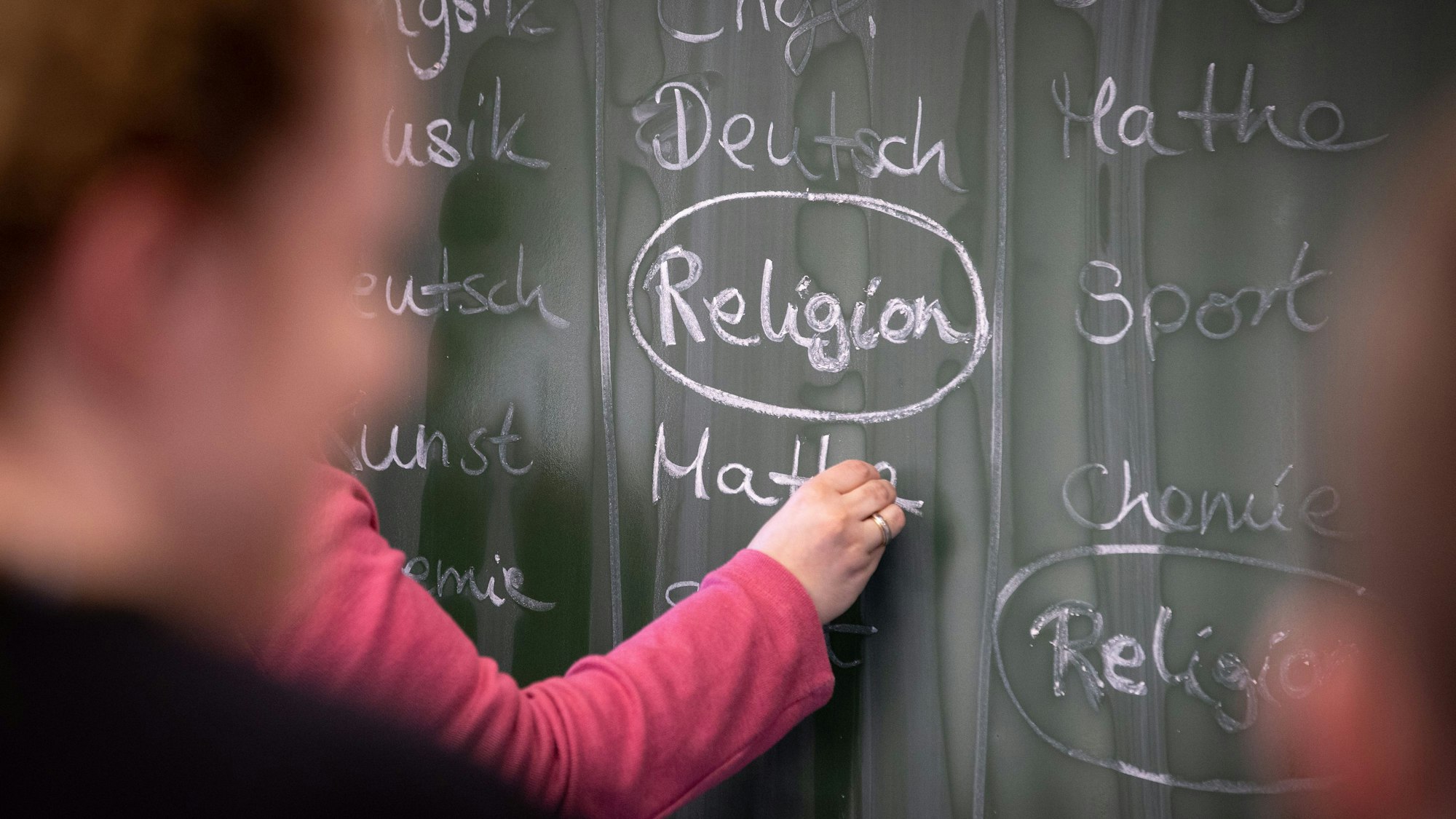 Eine Lehrerin schreibt mit Kreide einen Stundenplan mit Religionsunterricht an eine Tafel.
