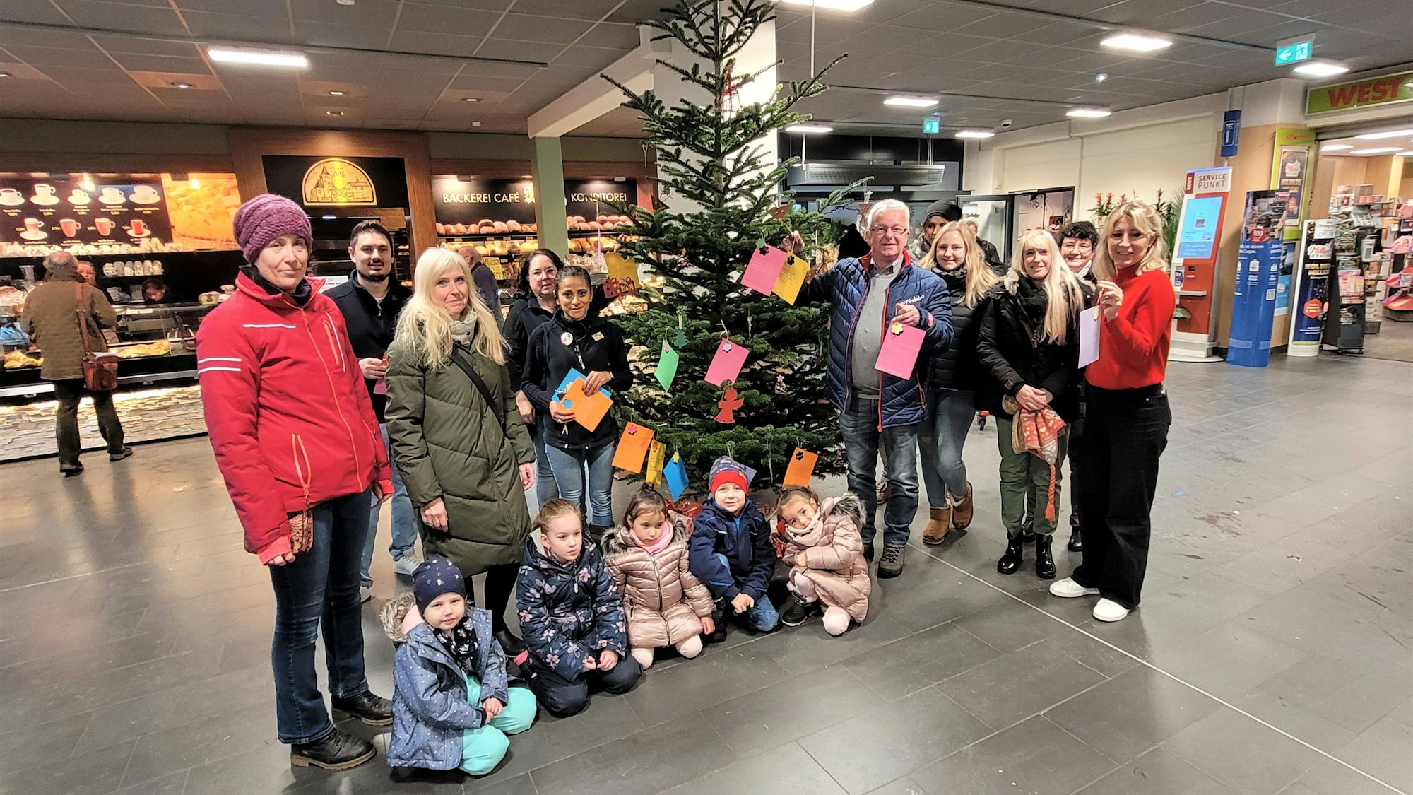 Frauen und Männer stehen im Kaller Rewe-Center an einem Weihnachtsbaum mit Wunschkarten und einige Kinder knien davor.