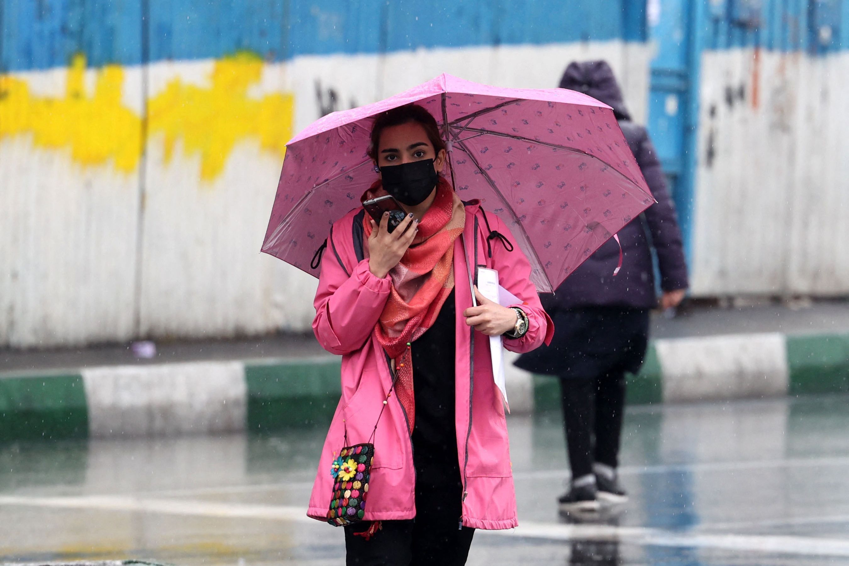 Eine junge iranische Frau läuft ohne Kopftuch an einem Regentag über die Straßen Teherans. Sie trägt einen Mund-Nasen-Schutz und hat einen rosafarbenen Regenschirm dabei.