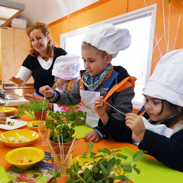 Eine Frau bereitet mit Kindergartenkindern, die Kochmützen tragen, Fruchtspieße zu.