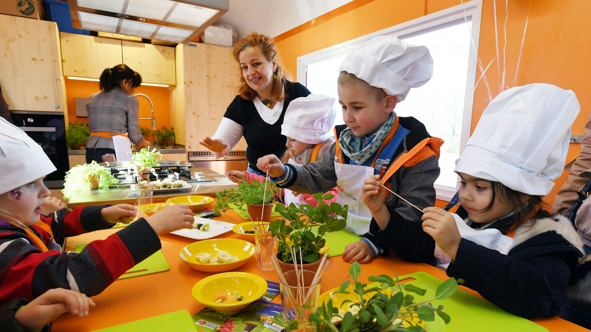 Eine Frau bereitet mit Kindergartenkindern, die Kochmützen tragen, Fruchtspieße zu.