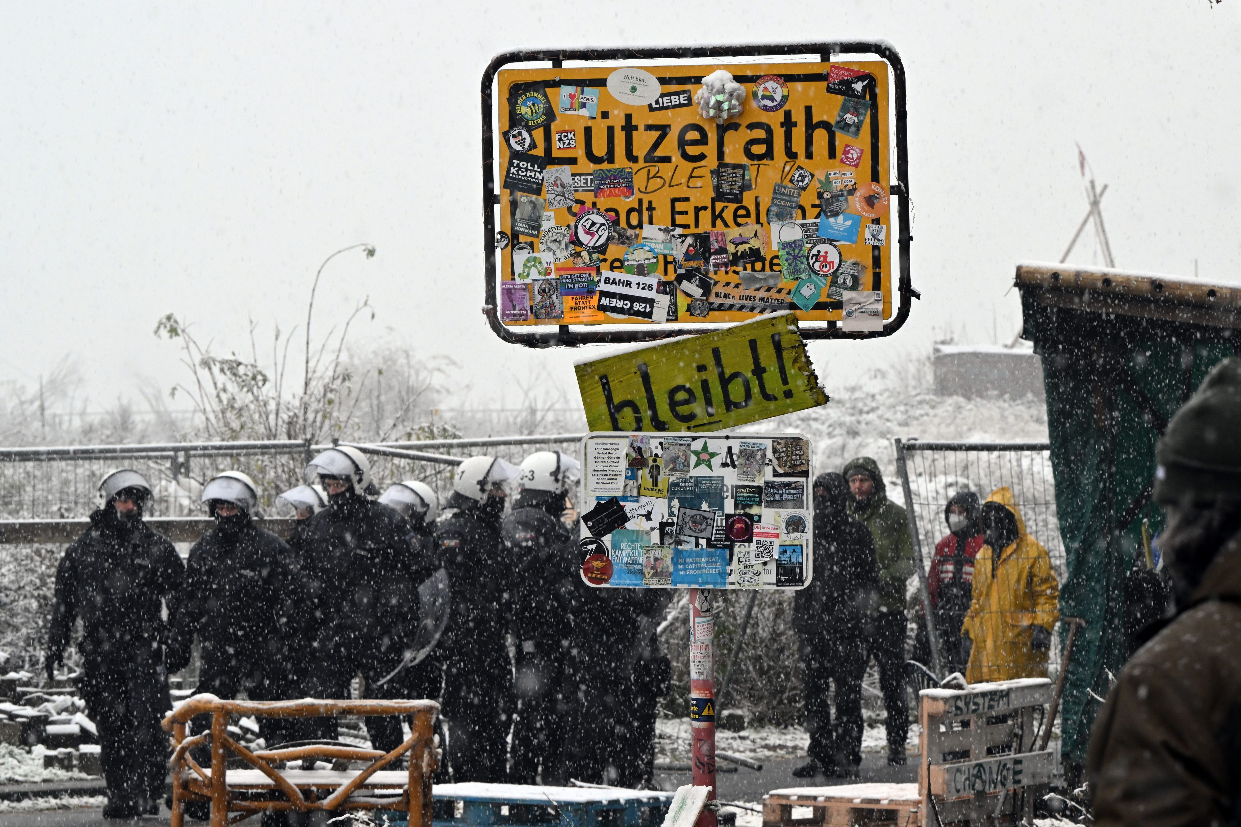 Die Polizei und Aktivisten der Initiative "Lützerath lebt!" stehen vor dem besetzten Gebiet hinter dem Ortsschild, während Schnee fällt.