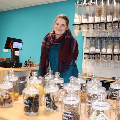 Eine Frau mit dunklem Haar steht hinter einem Verkaufstresen, auf dem Gläser mit Keksen und anderem zu sehen sind.